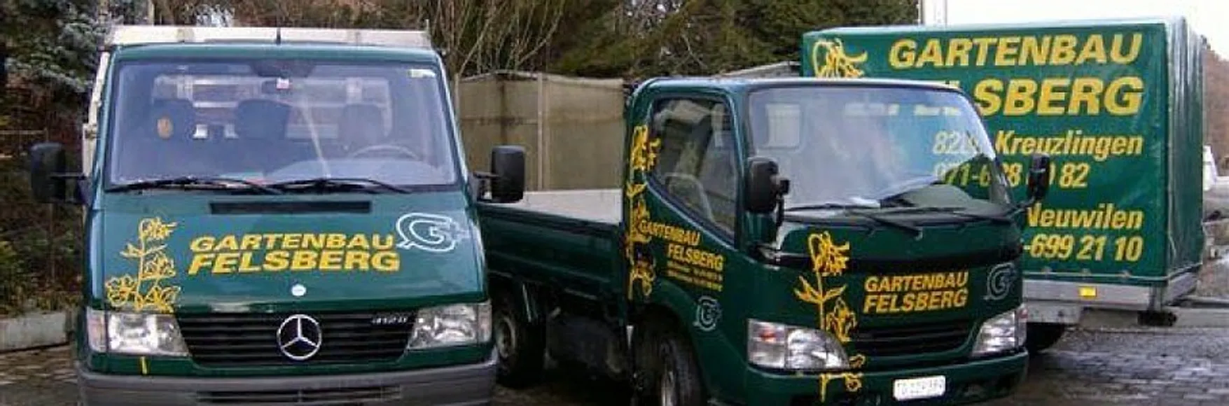Zwei Lastwagen und ein Anhänger auf denen das Firmenlogo von Gartenbau Felsberg abgedruckt sind